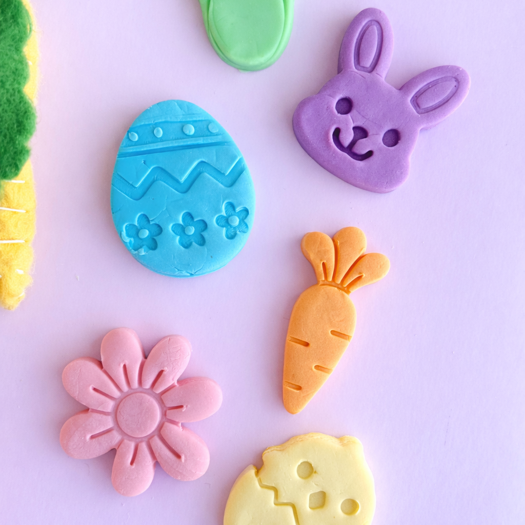 Colorful clay decorations shaped like a carrot, egg, flower, and other Easter-themed items on a light purple background.